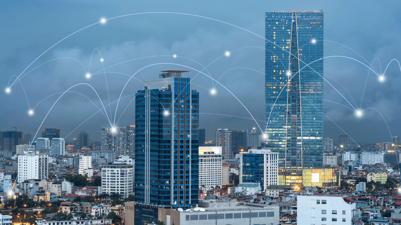 This image depicts a modern cityscape at dusk, with tall skyscrapers and various buildings illuminated by lights. Superimposed over the image are glowing points connected by curved lines, symbolizing a network or connectivity, representing a digital or telecommunications infrastructure. The connections seem to link different buildings, suggesting data flow, communication, or smart city technology. 